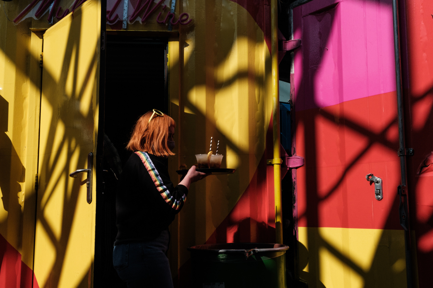 woman with red hair and stripes on sleeves carrying cocktails on a tray passing a very colourful hello, pink and red wall with strong shadows and shapes