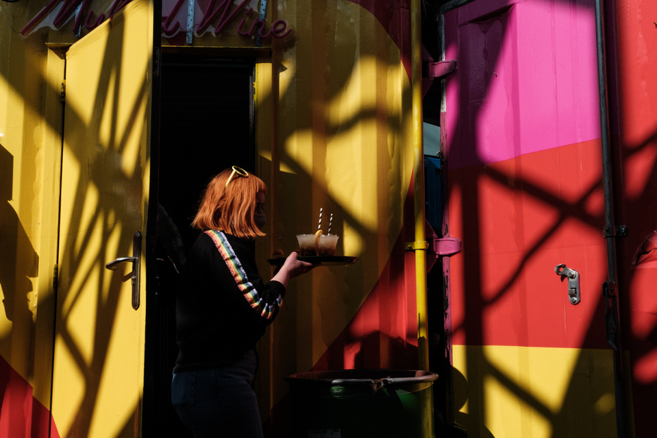 woman with orange hair carrying cocktails on a tray walks past a yellow, pink and red metal facade crisscrossed with bold black shadows