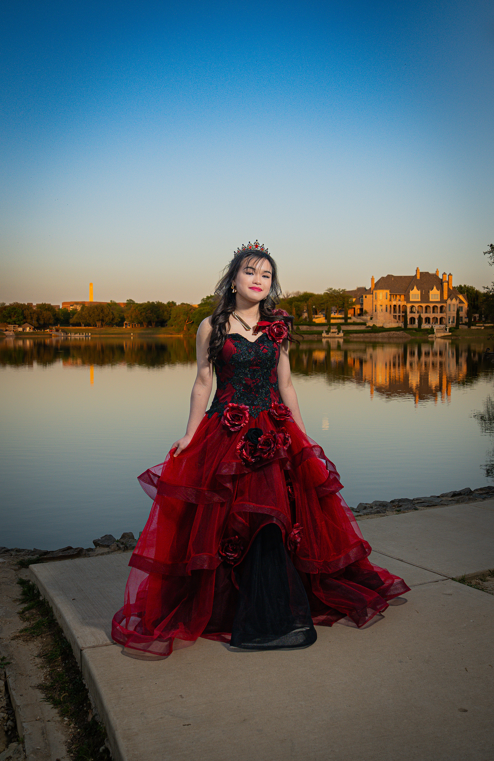 A debutante in a red and black gown stands tall beside the Adriatica Village lake, glowing under the golden-hour sun with soft reflections behind her.