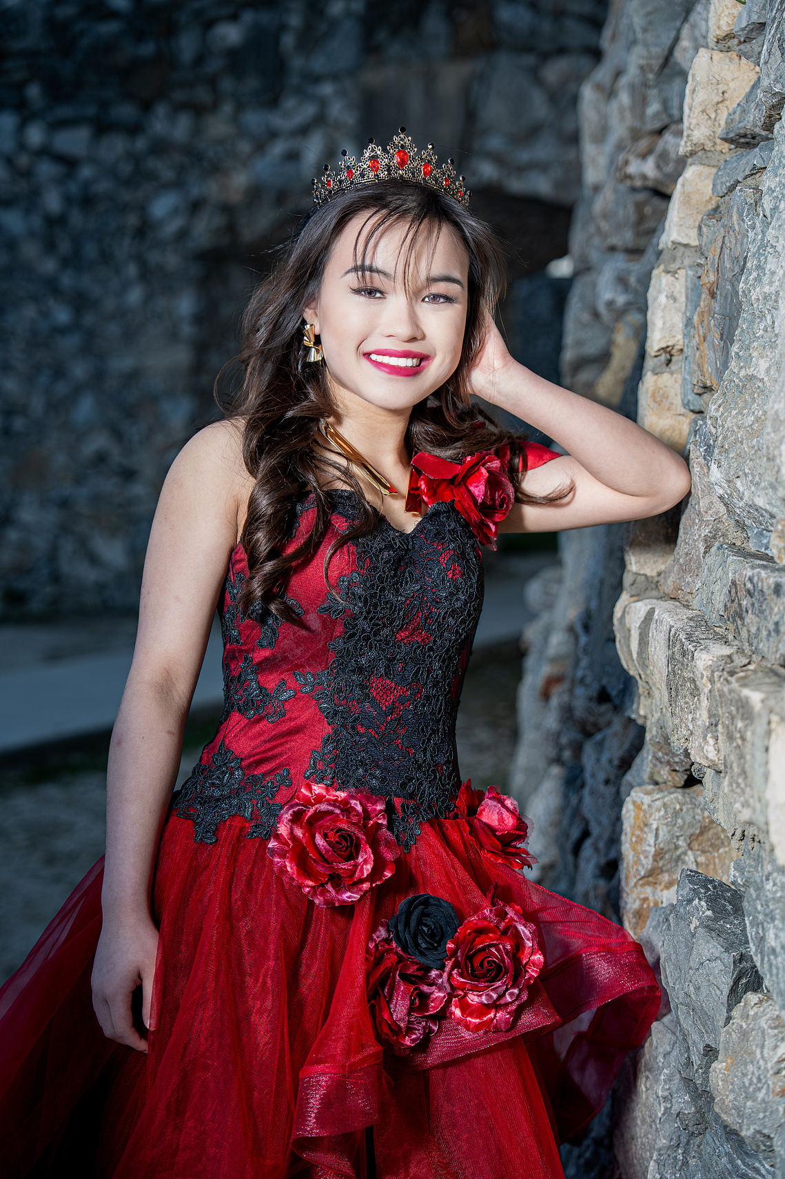 Young woman in a red and black floral gown smiling beside a rustic stone wall at Adriatica Village during her pre-debut photoshoot.