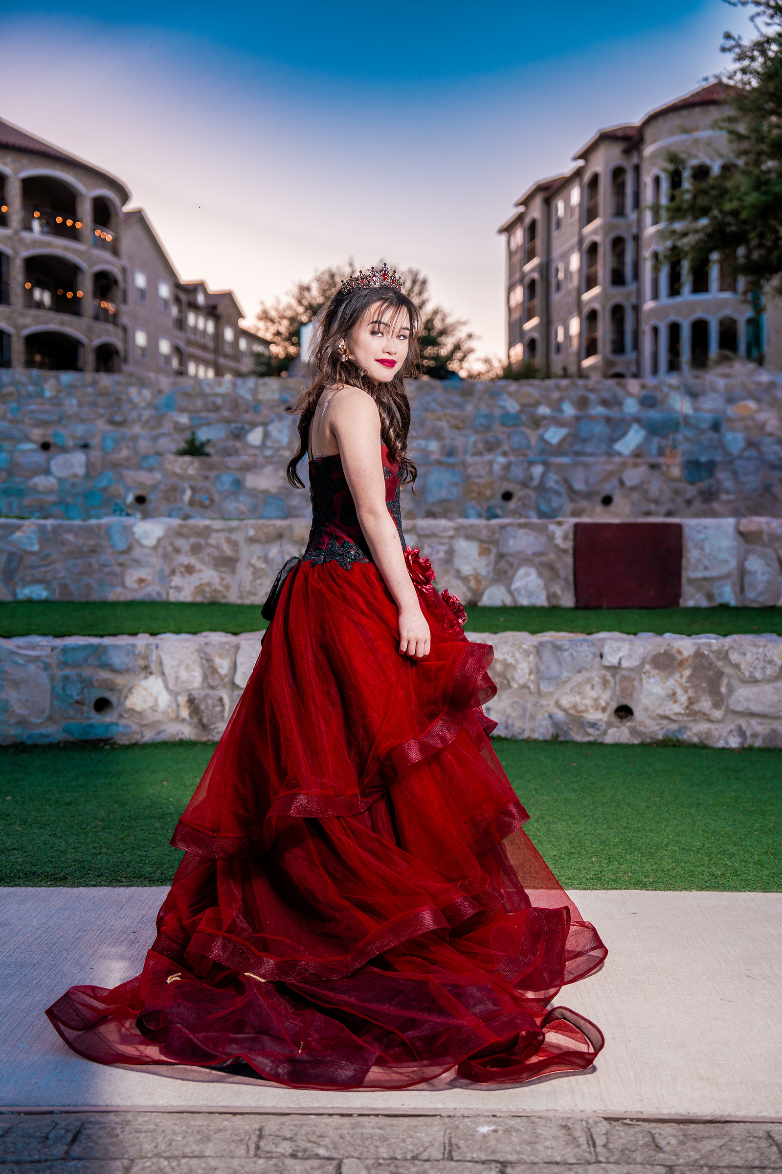 A full-length portrait of a debutante in a red gown looking over her shoulder, captured in front of modern stone architecture at Adriatica Village in McKinney, Texas.