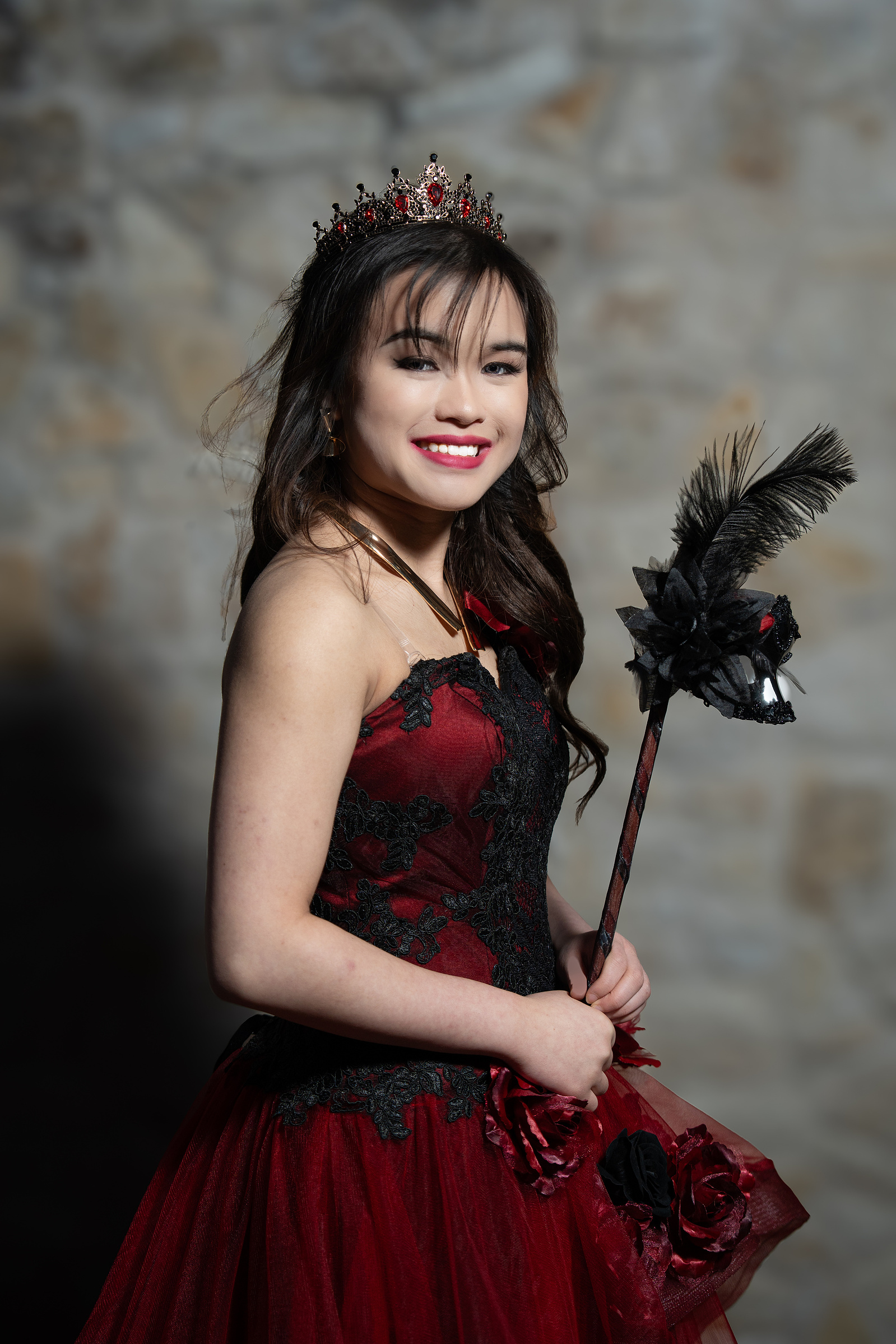 A close-up portrait of a young woman in a red gown holding a black floral prop, smiling confidently against a stone wall background in Adriatica Village.