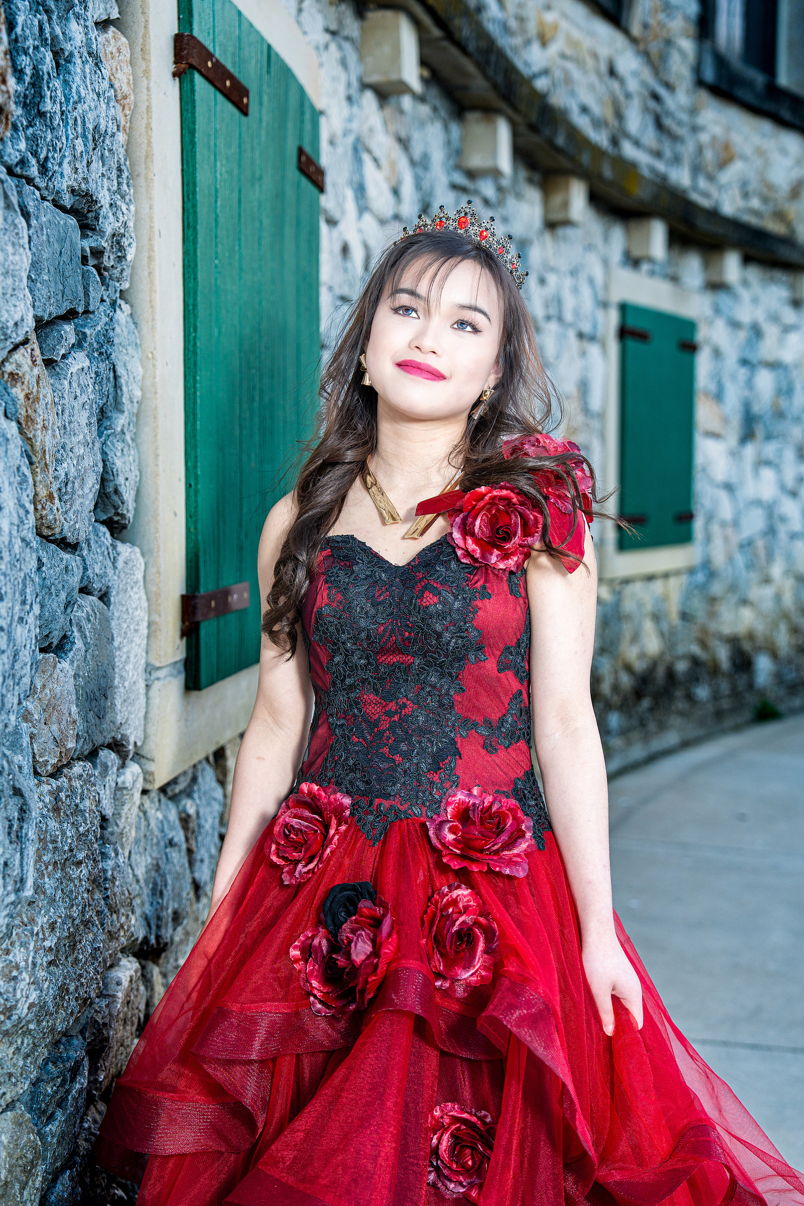 A close-up portrait of a debutante smiling while leaning on a stone wall at Adriatica Village, her red floral gown and confident pose adding modern elegance to the rustic setting.