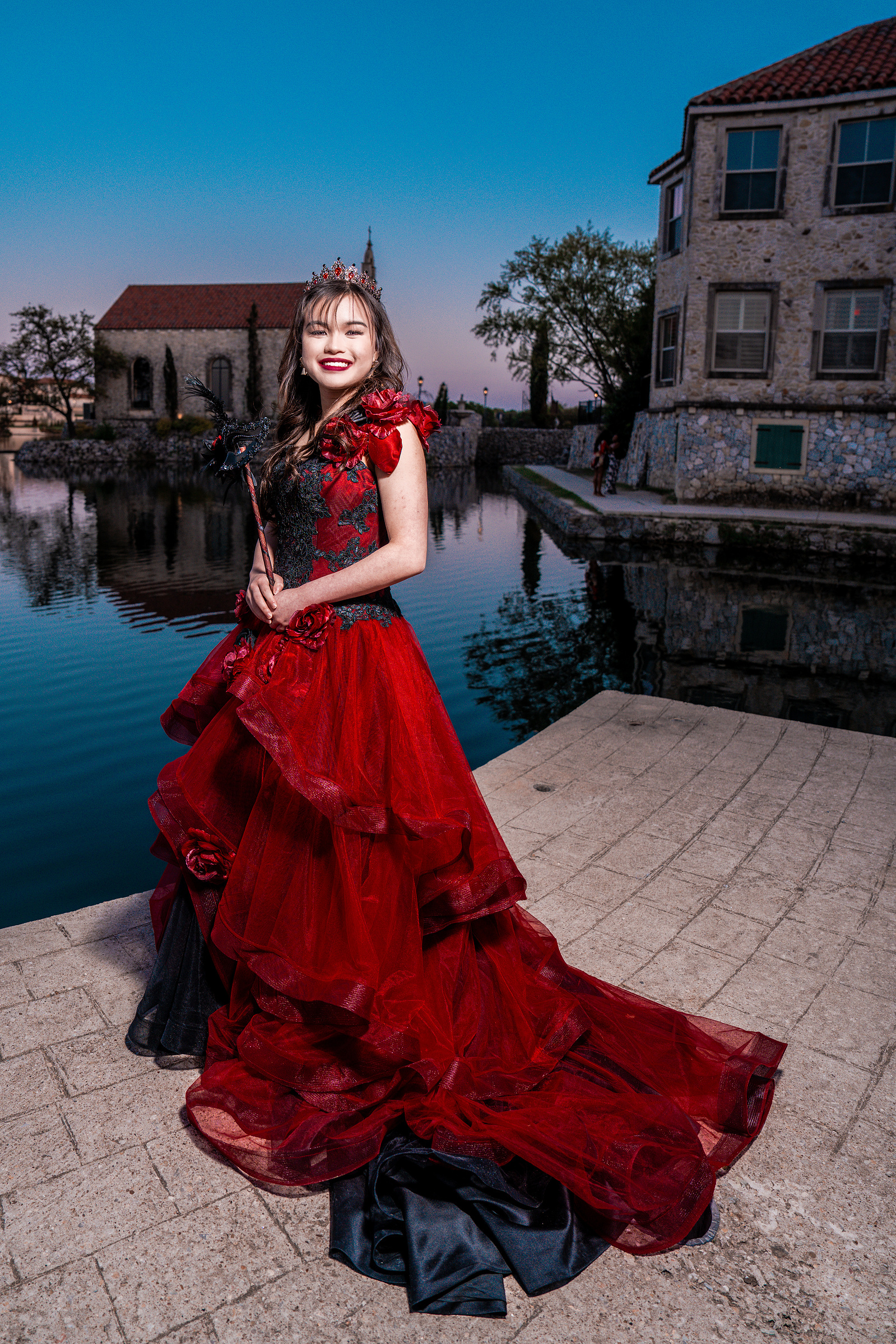A debutante in a flowing red ballgown stands near the stone edge of Adriatica Village’s lakefront, surrounded by rustic architecture and calm waters.