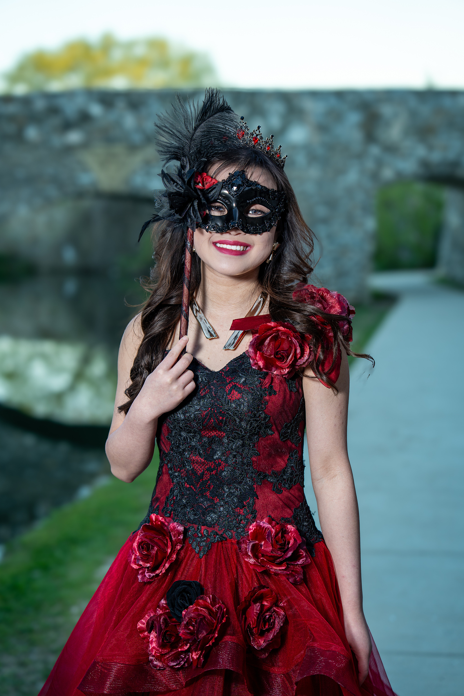 A close-up pre-debut portrait of a young woman in a red floral gown standing against a textured stone wall at Adriatica Village, showcasing her natural beauty and poise.