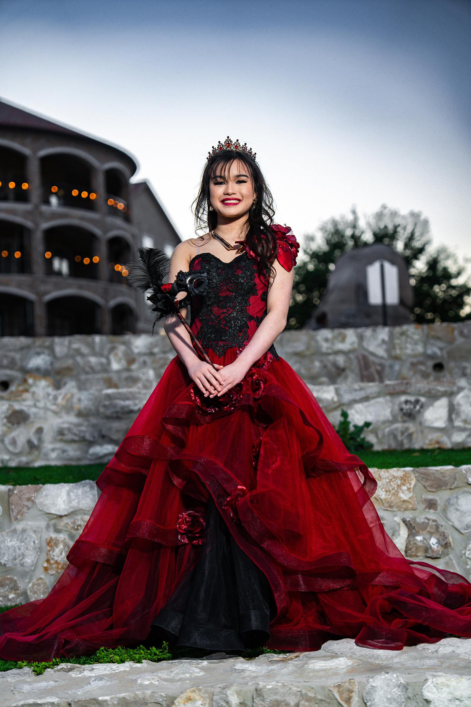 A debutante poses confidently in a red and black gown on a stone path at Adriatica Village, with a backdrop of rocky textures and soft greenery