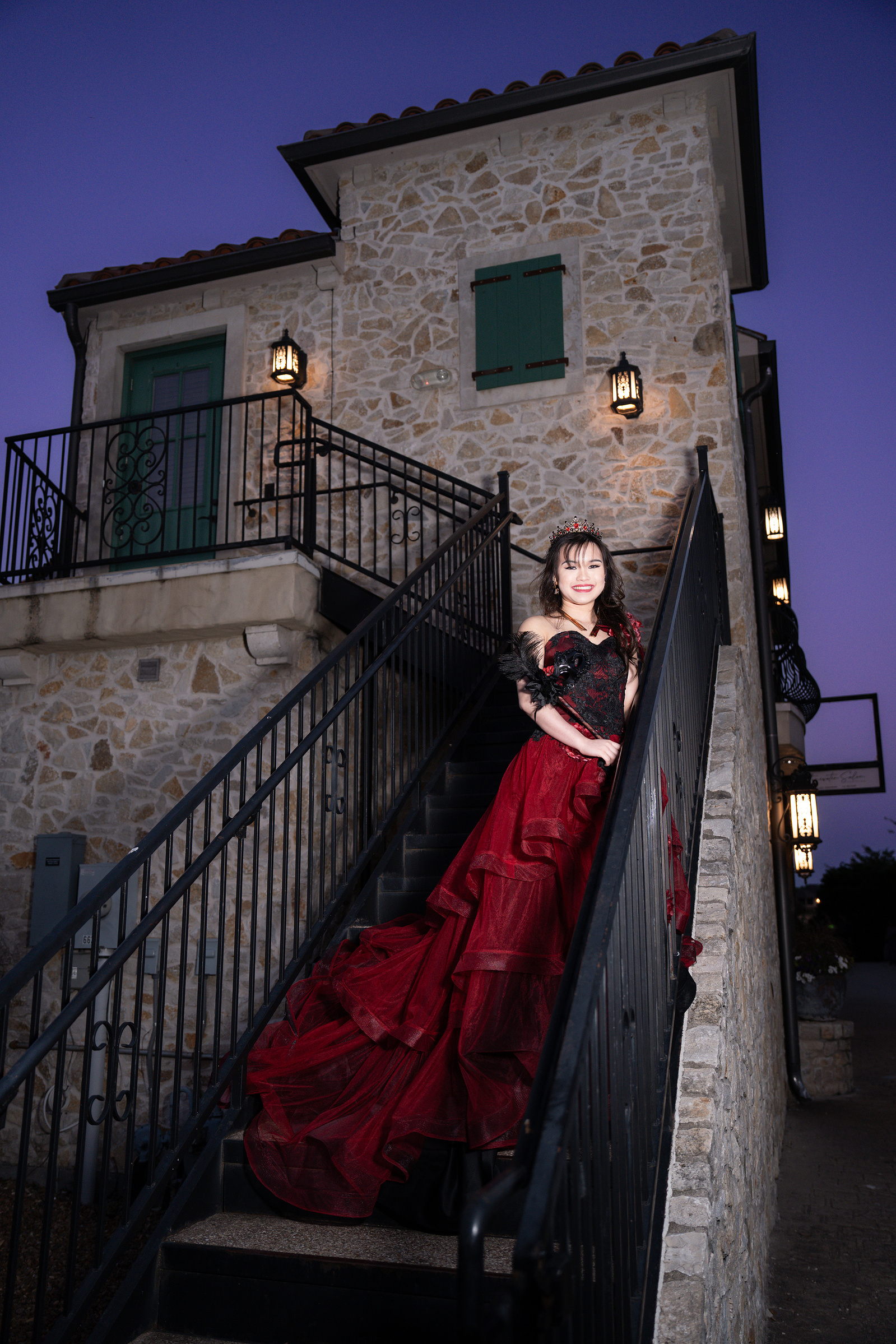 A debutante poses confidently on a staircase in Adriatica Village, her red gown cascading down the steps while evening light creates a cinematic mood.