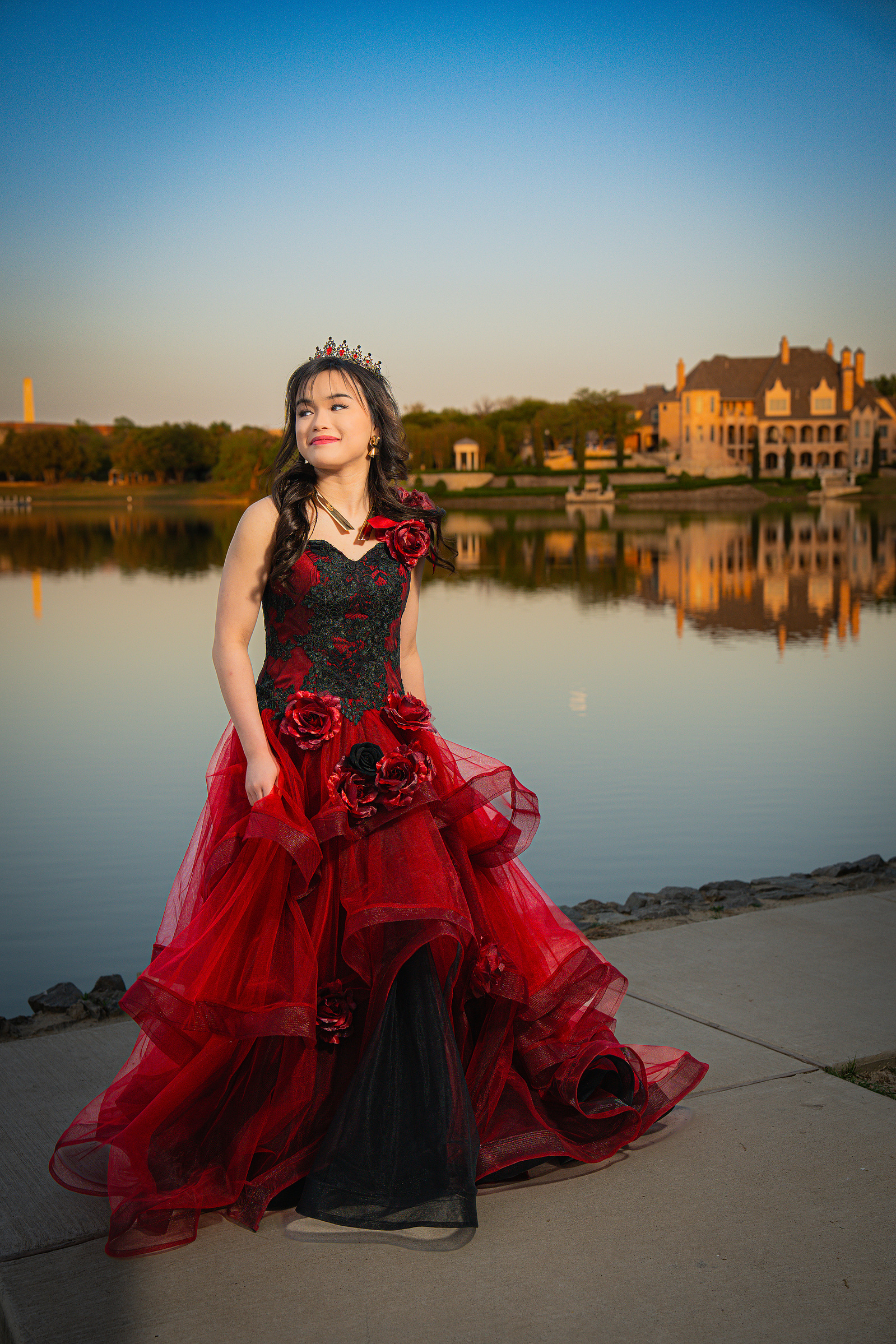 A young woman in a layered red dress poses along the lakefront at Adriatica Village, surrounded by soft water reflections and warm evening light.