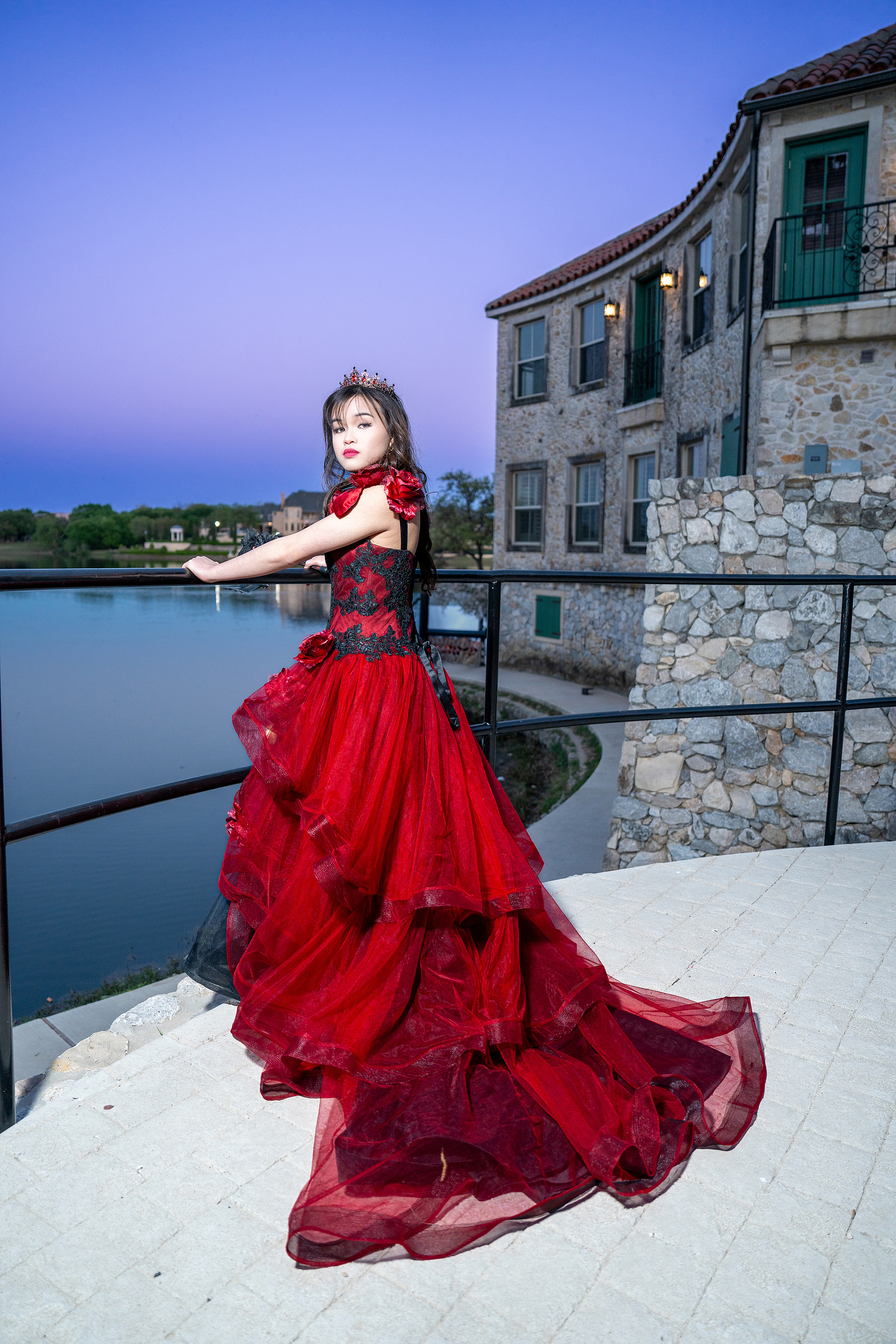 A young woman in a dramatic red ballgown leans against a black railing at Adriatica Village in McKinney, with stone buildings and golden hour light framing the scene.