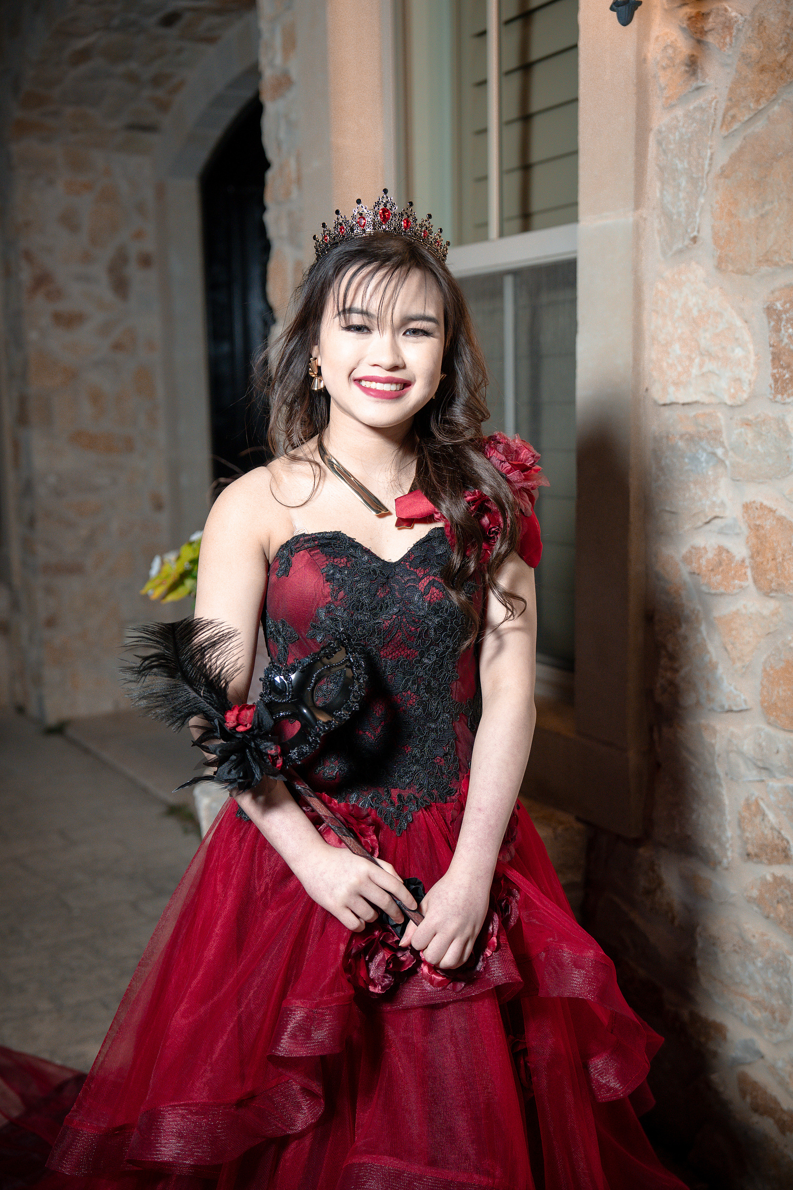 A joyful debutante smiles warmly in front of a stone pillar at Adriatica Village, her red dress and bouquet adding color to the neutral backdrop.