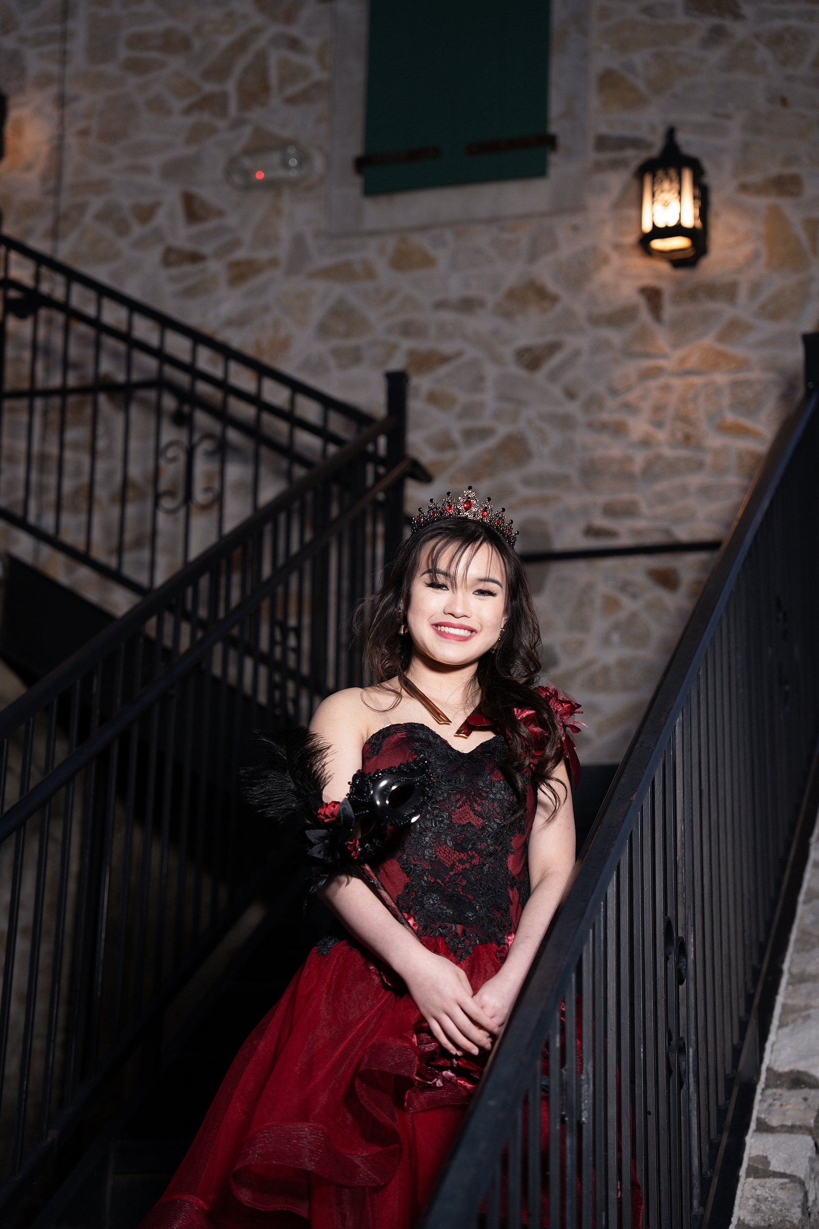 A pre-debut portrait of a debutante smiling on an outdoor staircase at Adriatica Village, with warm light highlighting her tiara and red floral gown.