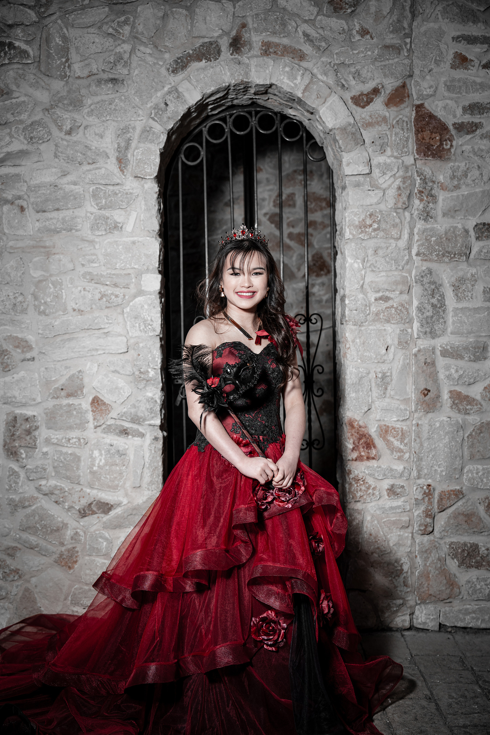 A regal portrait of a young woman standing in front of a textured stone arch, holding a floral prop while wearing a detailed red and black debutante gown.