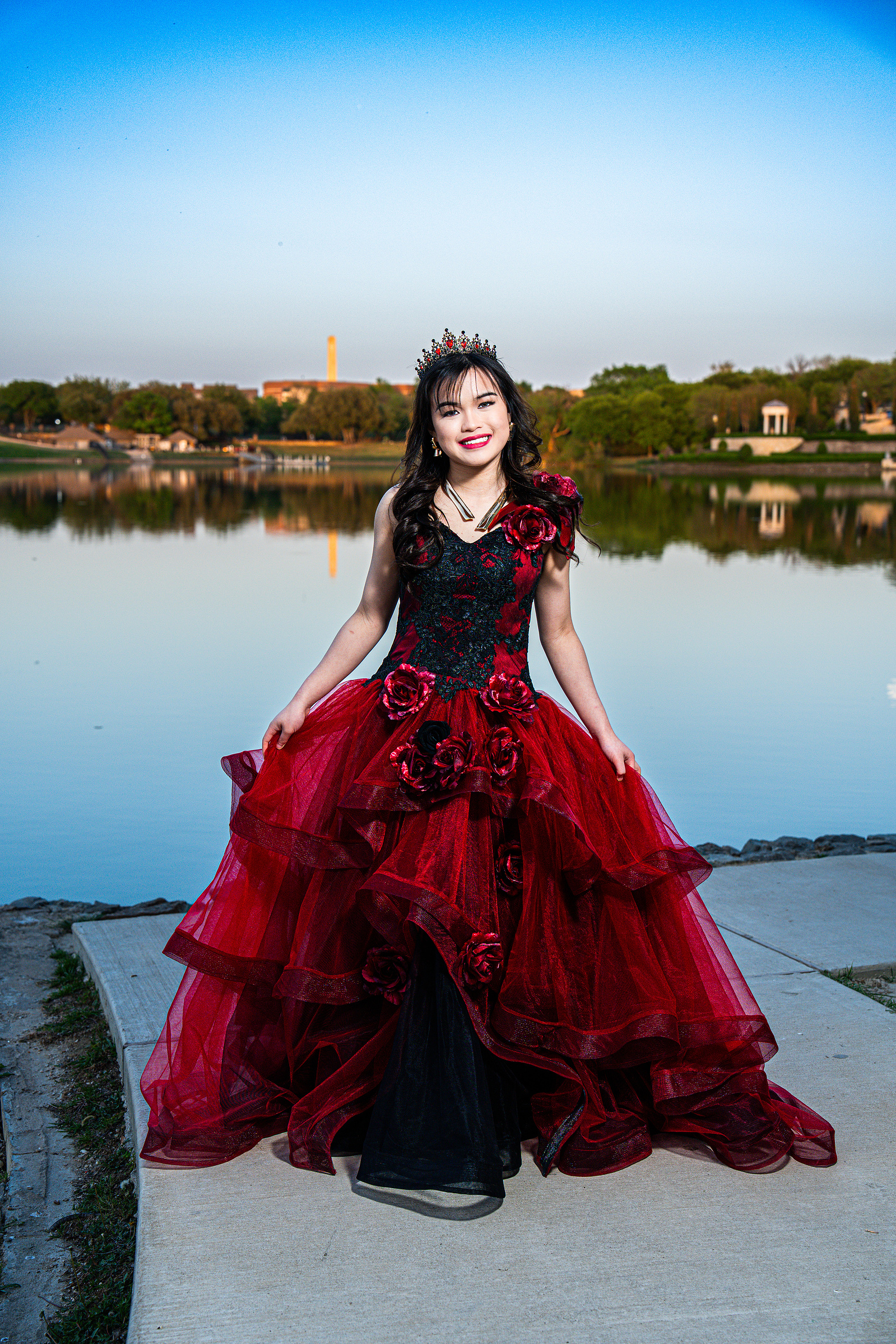 A regal debutante in a deep red gown stands against a vibrant lakefront backdrop at Adriatica Village, framed by lush trees and serene waters.
