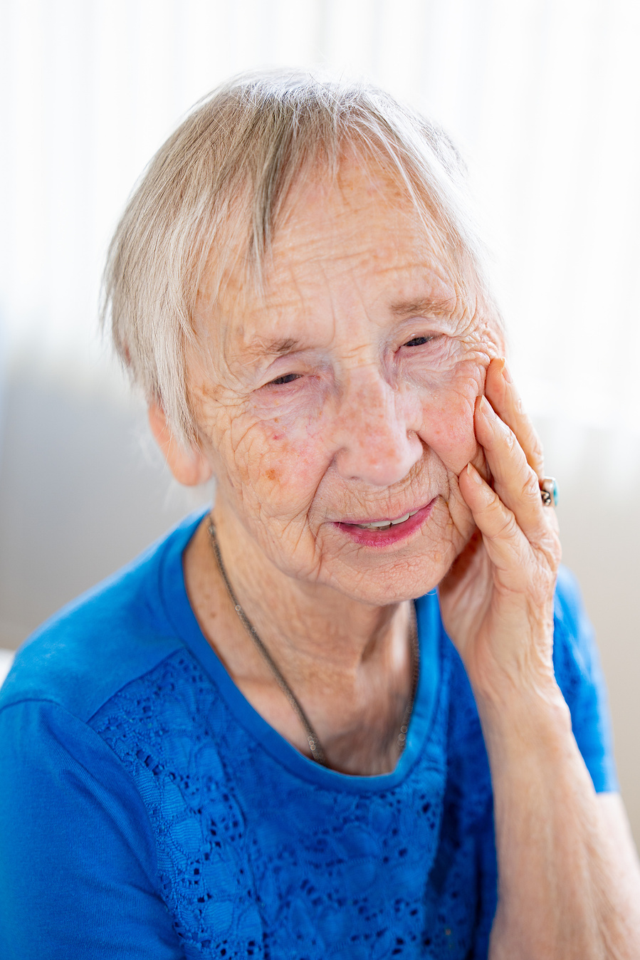 A joyful 90-year-old woman smiles with her hand gently on her cheek, capturing the grace and elegance of aging in a natural light senior portrait.