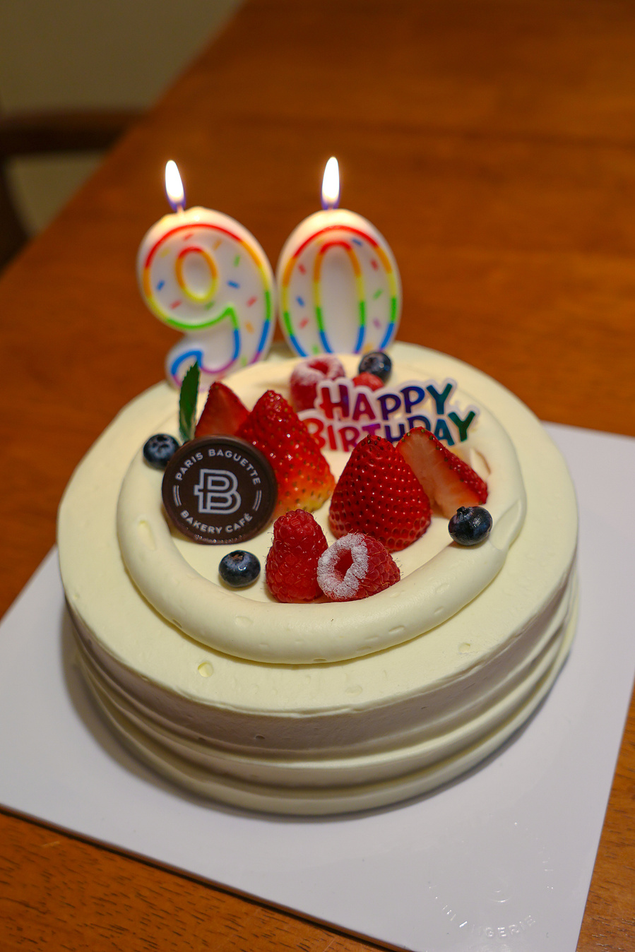 A cheerful elderly woman in a crown holds her birthday cake with joy, capturing the spirit of turning 90 with laughter and love in a casual home setting.