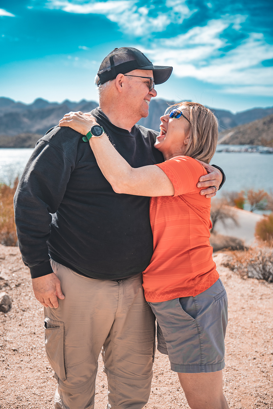 Smiling couple standing by the shoreline at Saguaro Lake with blue skies and mountain views in the background.