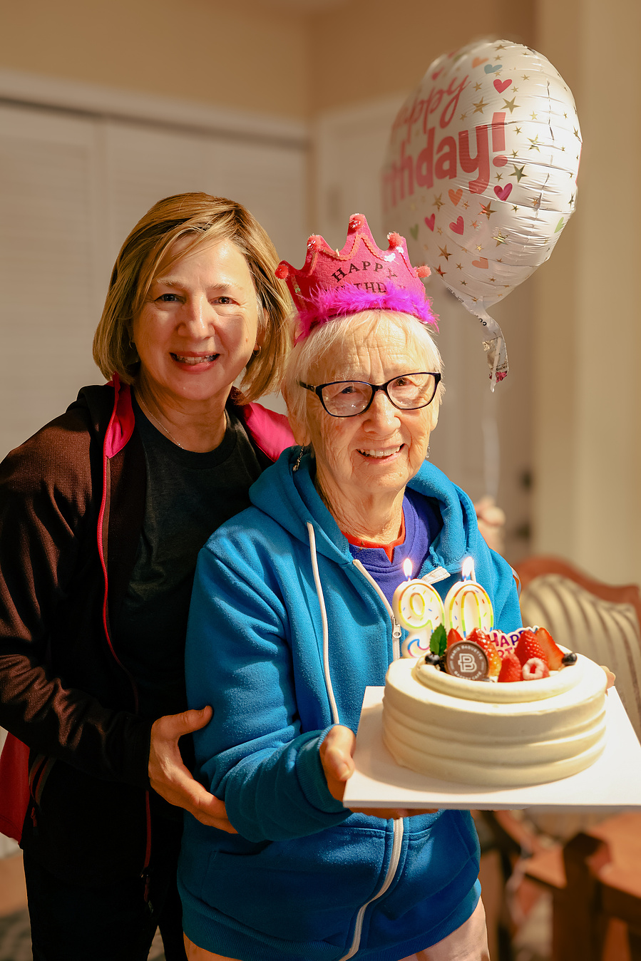 This cheerful birthday moment features a 90-year-old woman in a pink crown holding her cake with her daughter beside her, celebrating joyful aging and family milestones in a fun and festive portrait.