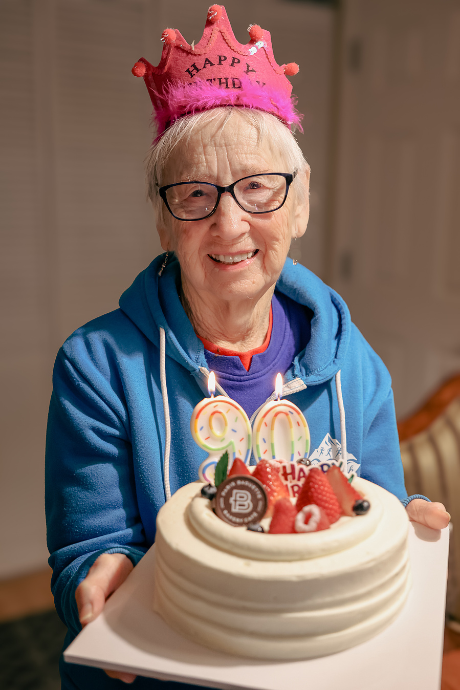 This senior birthday portrait features a smiling 90-year-old woman in glasses and a party crown, highlighting confidence and joy in aging gracefully.