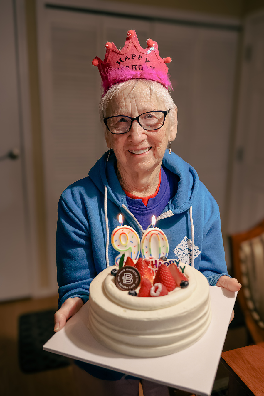A 90-year-old woman celebrates her birthday with a cake and balloon, wearing a pink crown and smiling proudly in this fun senior milestone portrait.