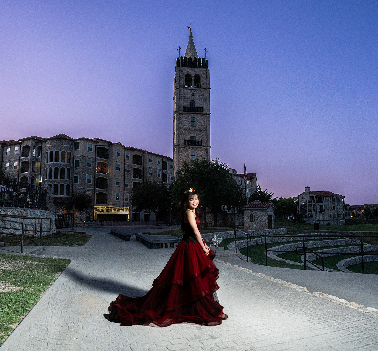 A dramatic twilight portrait of a debutante in a red gown at Adriatica Village, with the iconic bell tower and European-style buildings behind her.