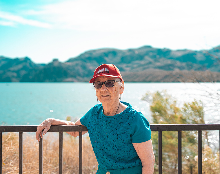 An active senior woman poses confidently by the lake in Arizona, wearing a red cap and sunglasses with scenic mountains and water in the background. This vibrant travel portrait highlights the joy of senior outdoor adventures, nature walks, and active age