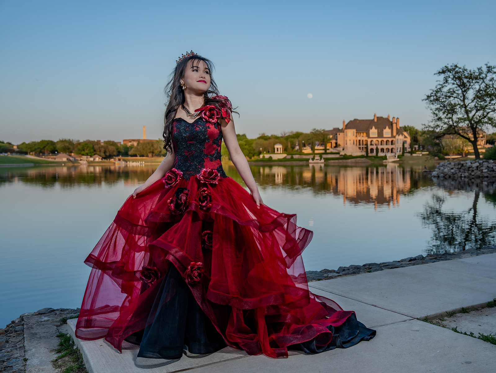 A young woman in a flowing red ballgown twirls playfully near the edge of the lake at Adriatica Village in McKinney, Texas, with golden-hour reflections on the water behind her.