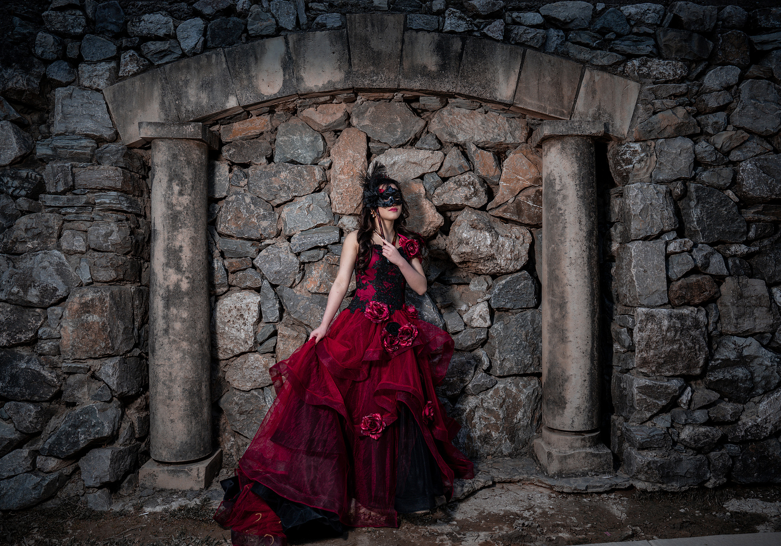 A debutante in a voluminous red gown sits gracefully between stone columns at Adriatica Village in McKinney, creating a dramatic, fairytale-inspired portrait.