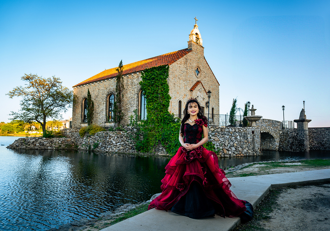 A young woman in a red pre-debut gown poses in front of Bella Donna Chapel at Adriatica Village in McKinney, Texas during golden hour. The European-inspired stone architecture and serene waterfront create a timeless fairytale backdrop for her milestone.