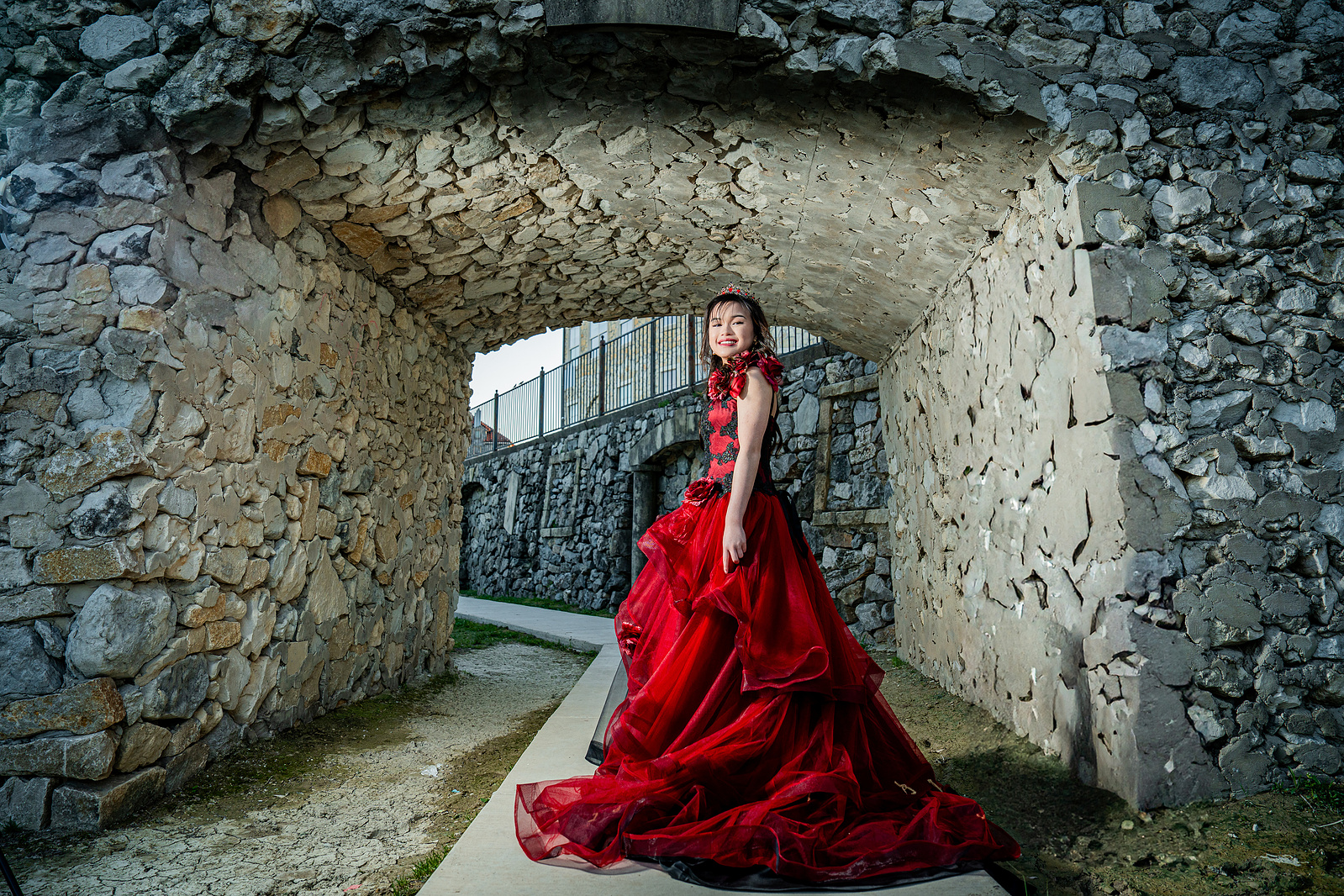 A young woman in a red ballgown poses mid-step under a charming stone archway at Adriatica Village in McKinney, Texas. The natural light highlights her flowing gown and joyful expression, capturing her elegant debut moment.