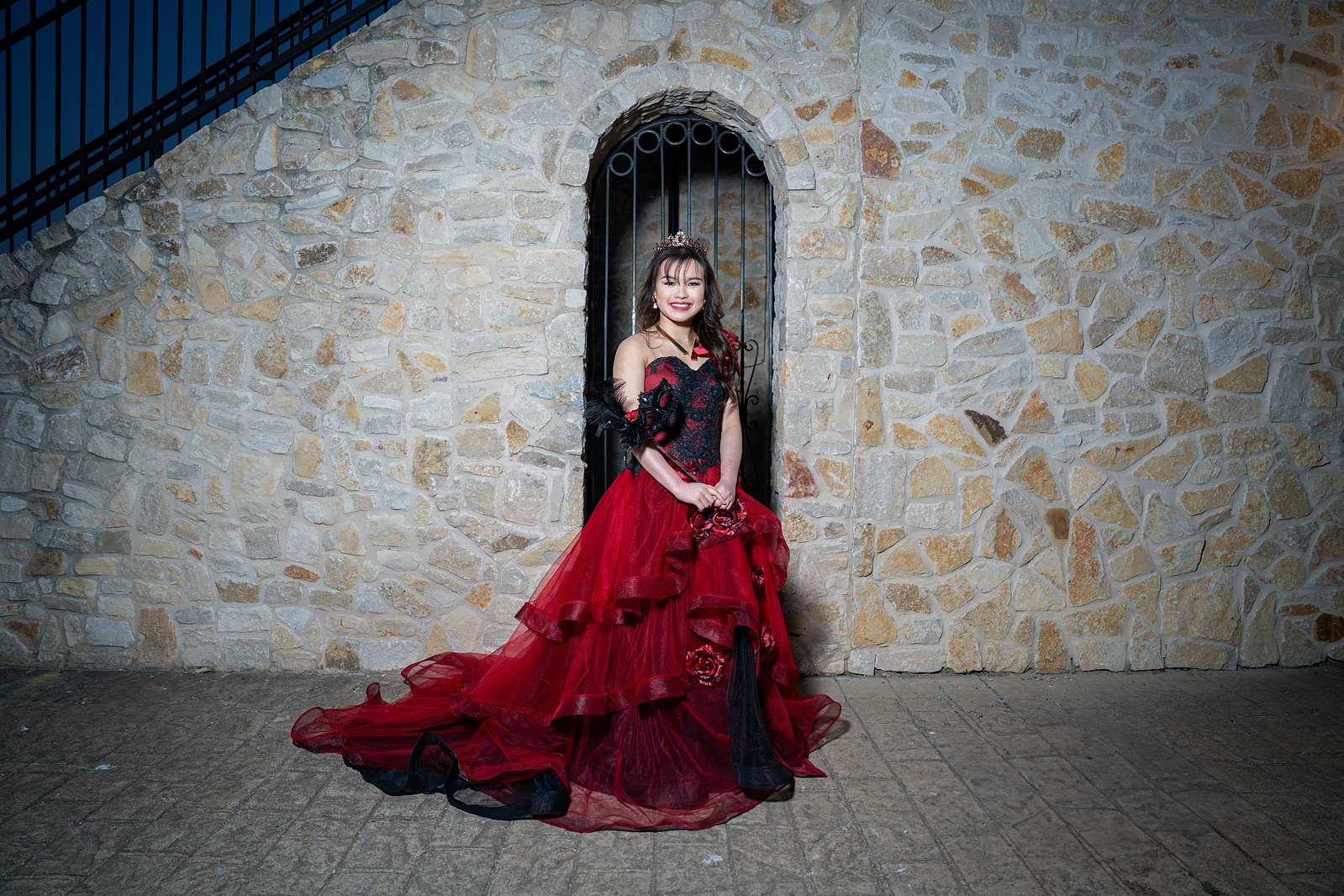 A debutante in a red gown sits in front of a rustic arched stone doorway at Adriatica Village, blending poise and elegance with the medieval-style setting.
