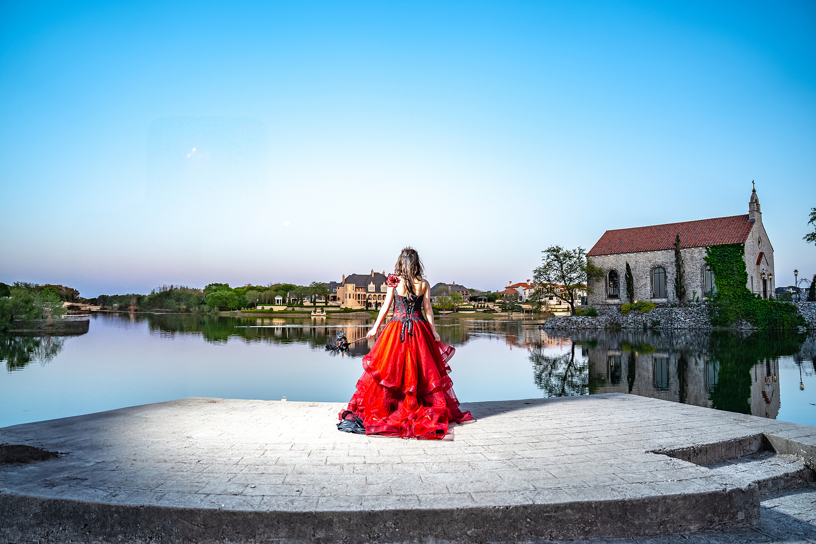 A debutante in a red gown stands centered on a white circular stone platform at Adriatica Village, McKinney, with blue skies and rustic European-style buildings in the background.