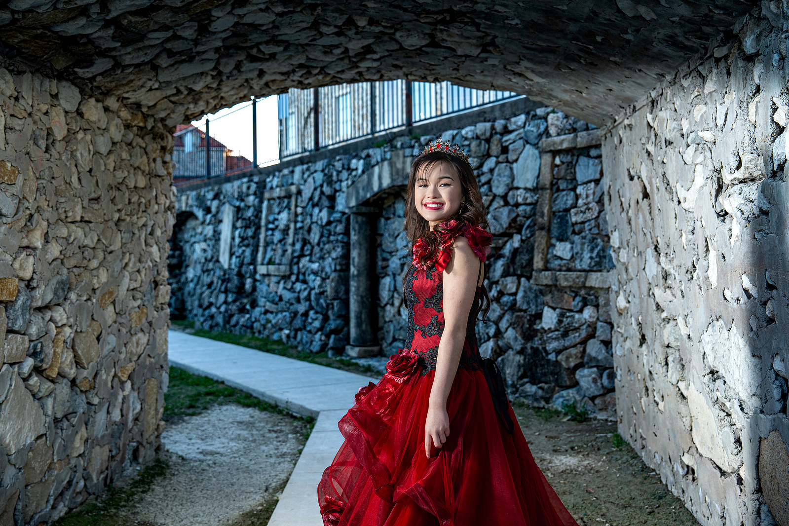 A debutante in a dramatic red gown sits gracefully beneath a rustic stone archway in Adriatica Village, McKinney. The tunnel’s textured backdrop frames her like a princess in a storybook setting.