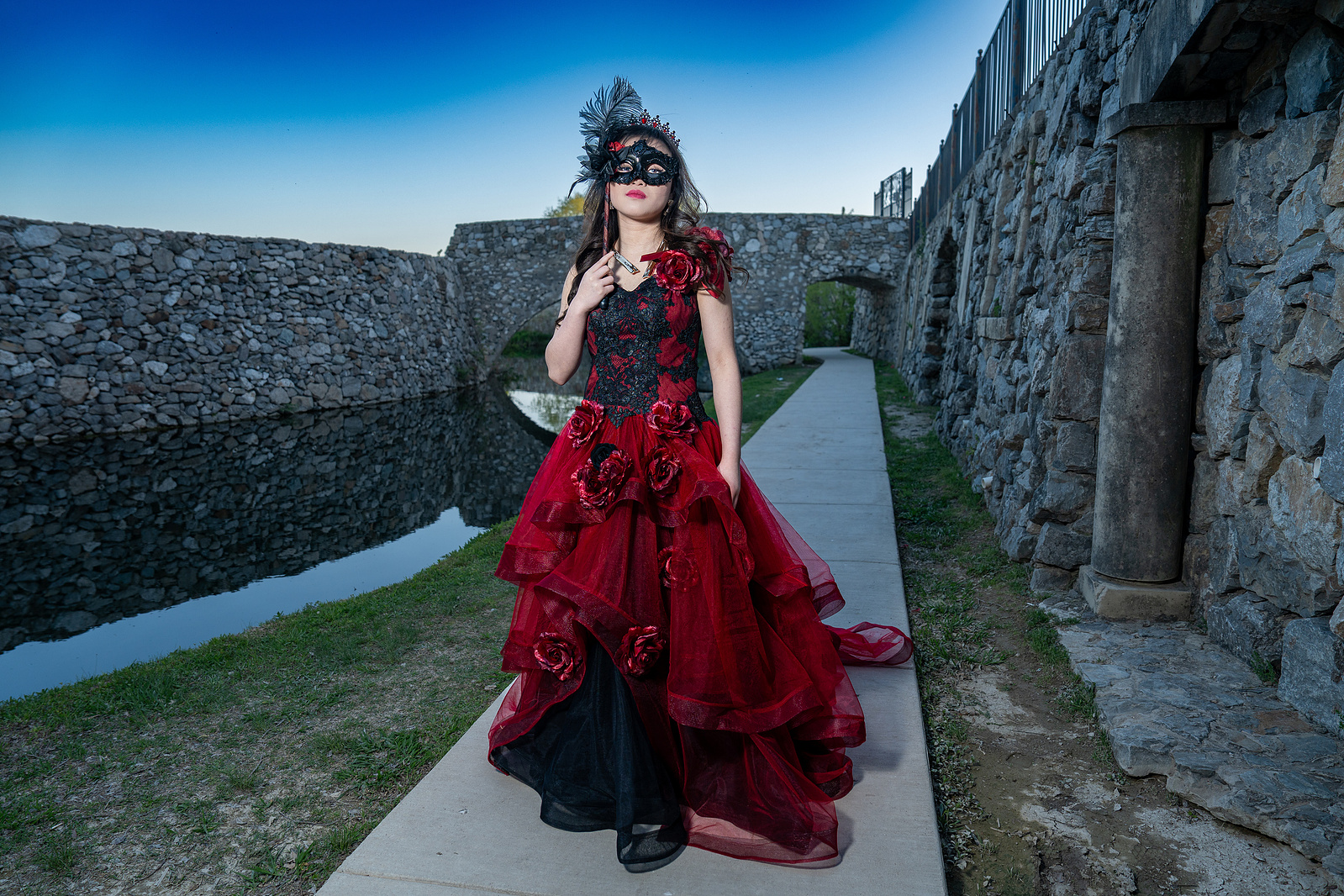 A debutante walks confidently in a red ballgown and black masquerade mask along a stone path at Adriatica Village in McKinney, Texas, evoking a mysterious, regal presence.