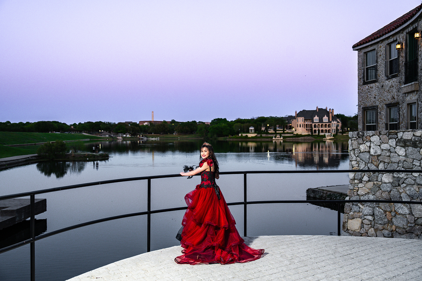 A debutante in a red gown poses near a railing overlooking the Adriatica Village lake at twilight, with soft purple skies and calm waters behind her.