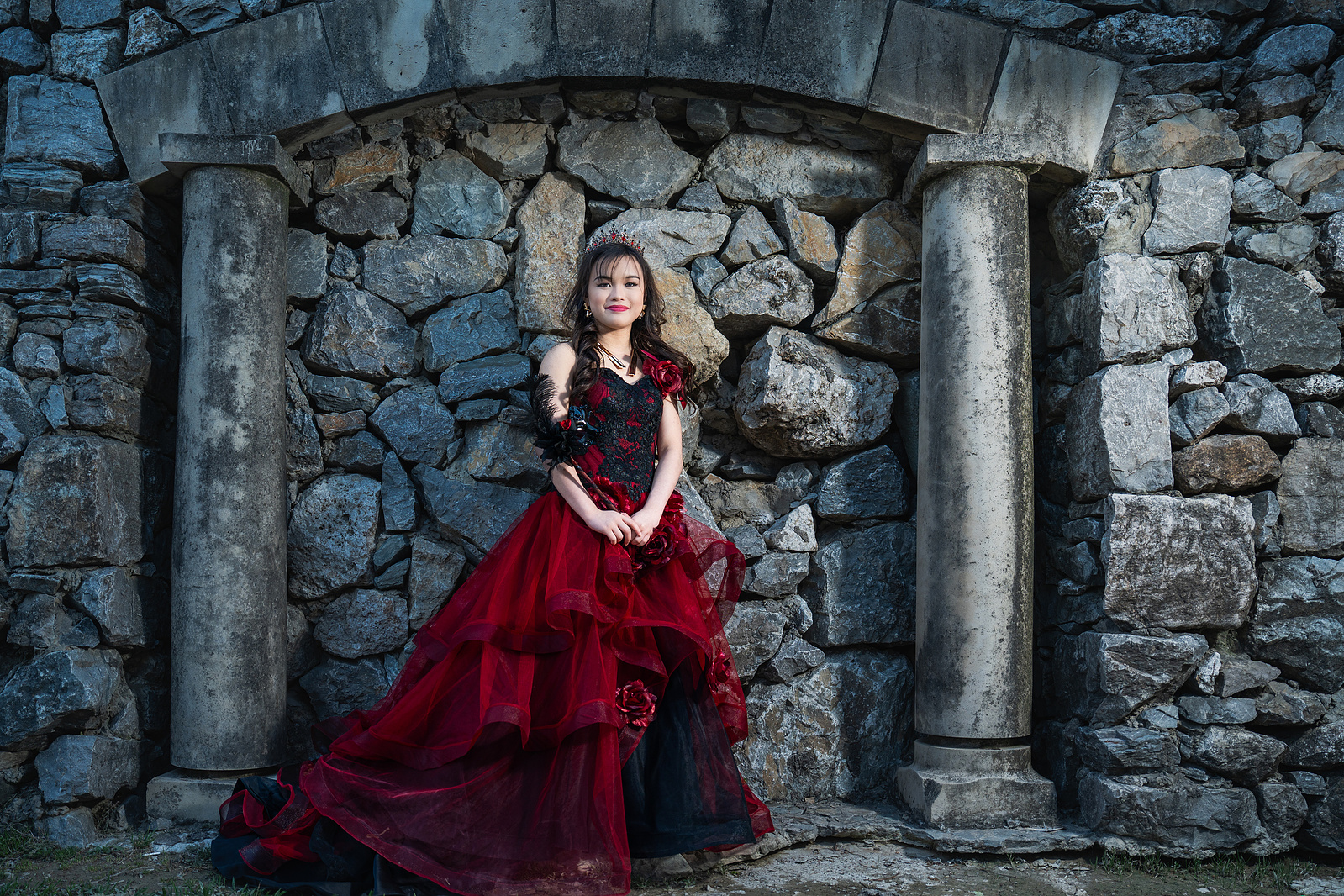 A young woman in a red and black debut gown and feathered masquerade mask smiles brightly in front of a stone wall at Adriatica Village, adding a playful, elegant touch to her pre-debut shoot.