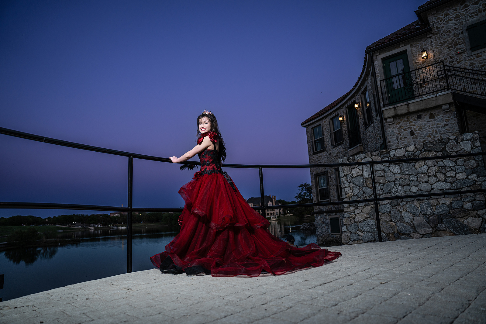 Captured at dusk, a young woman in a red gown walks gracefully beside the waterfront at Adriatica Village, surrounded by stone buildings and soft blue-purple skies.