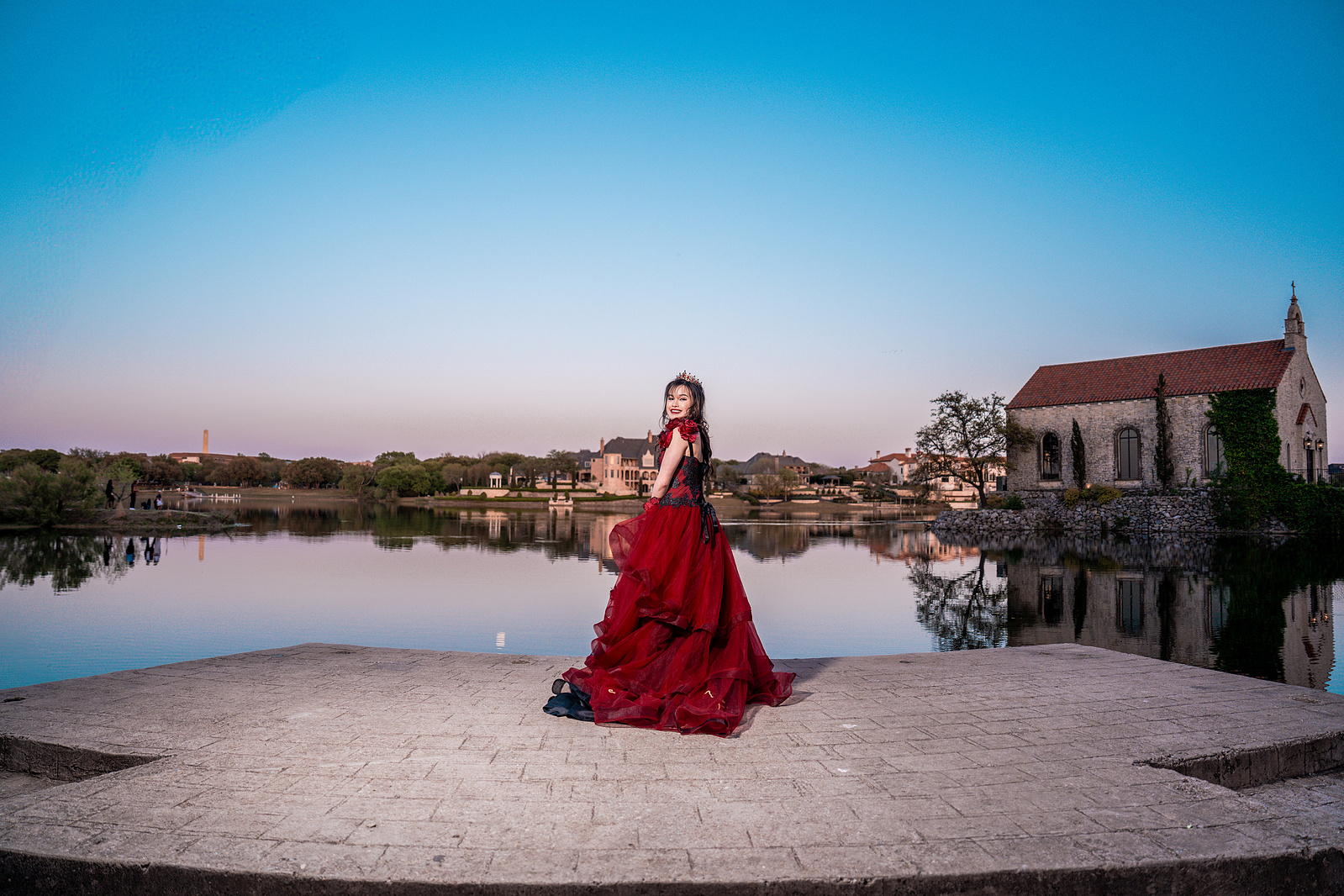 A young woman in a red dress stands alone on a white circular stage beside the Adriatica Village lake, exuding grace and poise during her pre-debut shoot.