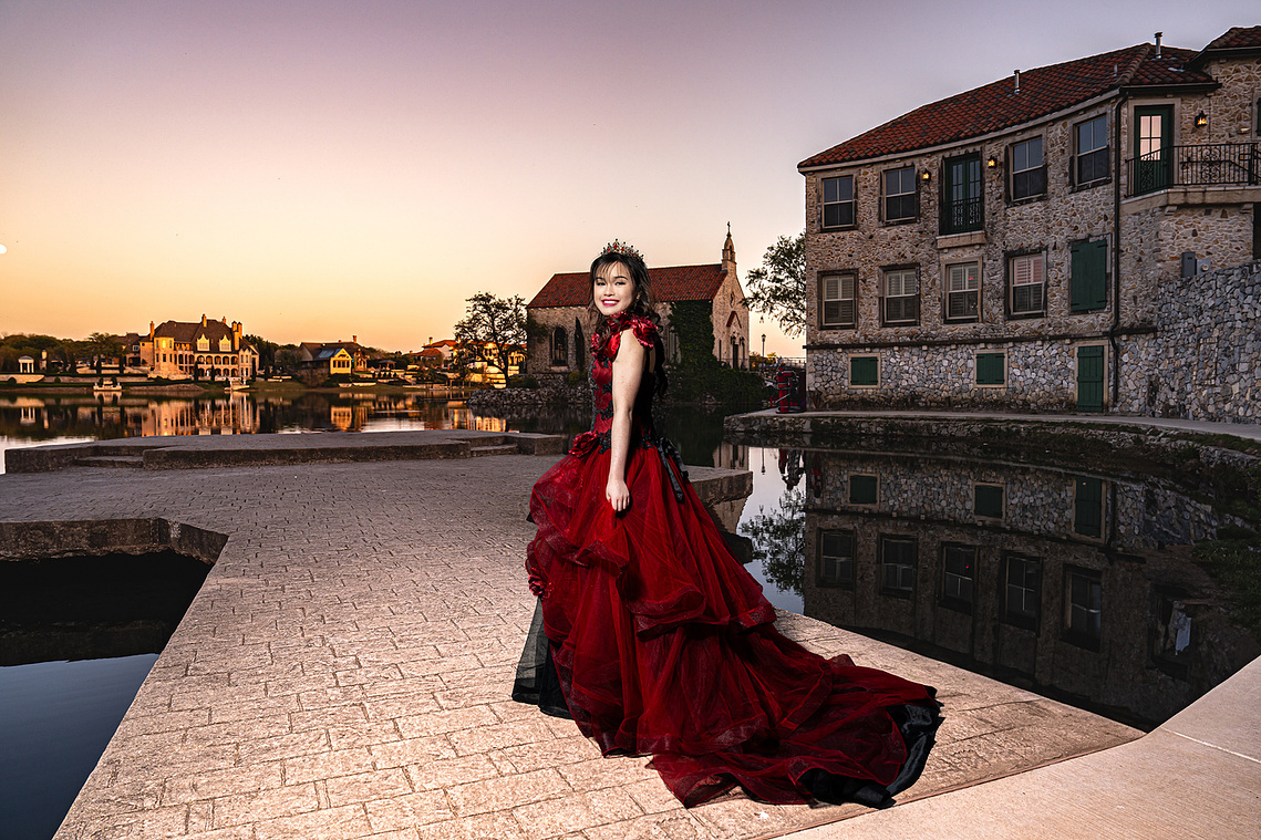 A young debutante in a dramatic red ballgown and tiara smiles as she poses at sunset in Adriatica Village, McKinney, Texas. The European-style stone buildings, still water reflections, and golden sky create a fairytale-like atmosphere perfect for an elega