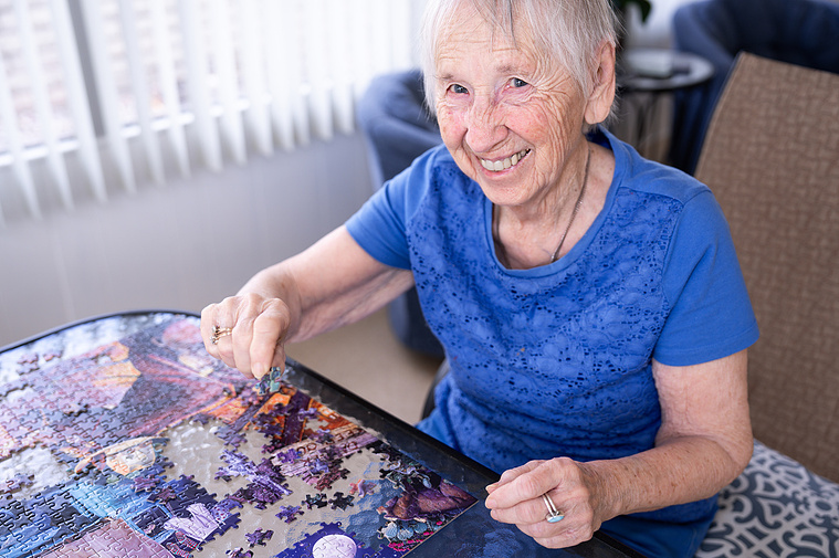 A smiling 90-year-old woman works on a colorful jigsaw puzzle in a bright, sunlit room. This candid portrait captures the joy of staying mentally active and engaged through hobbies like puzzles — perfect for lifestyle photography focused on senior wellnes