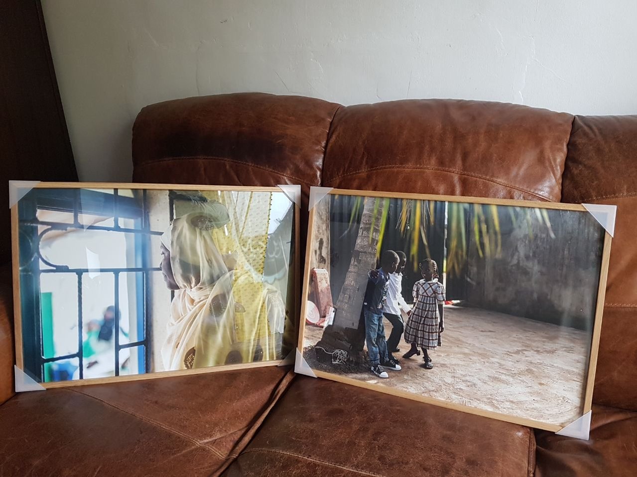 Two framed A2 prints, beside each other on a leather sofa.  The left picture shows a head and shoulders picture of a woman wearing a pale yellow and gold hijab, and looking out of a barred window. The right picture is  of 3 children stood in a courtyard.