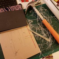 the text block, with black end paper and a piece of ankara stuck to the spine, on a cutting mat covered in paper trimmings that have been cut off with the scalpel that's on top of them.  also two metal rulers and a hammer that I used to round the spine.