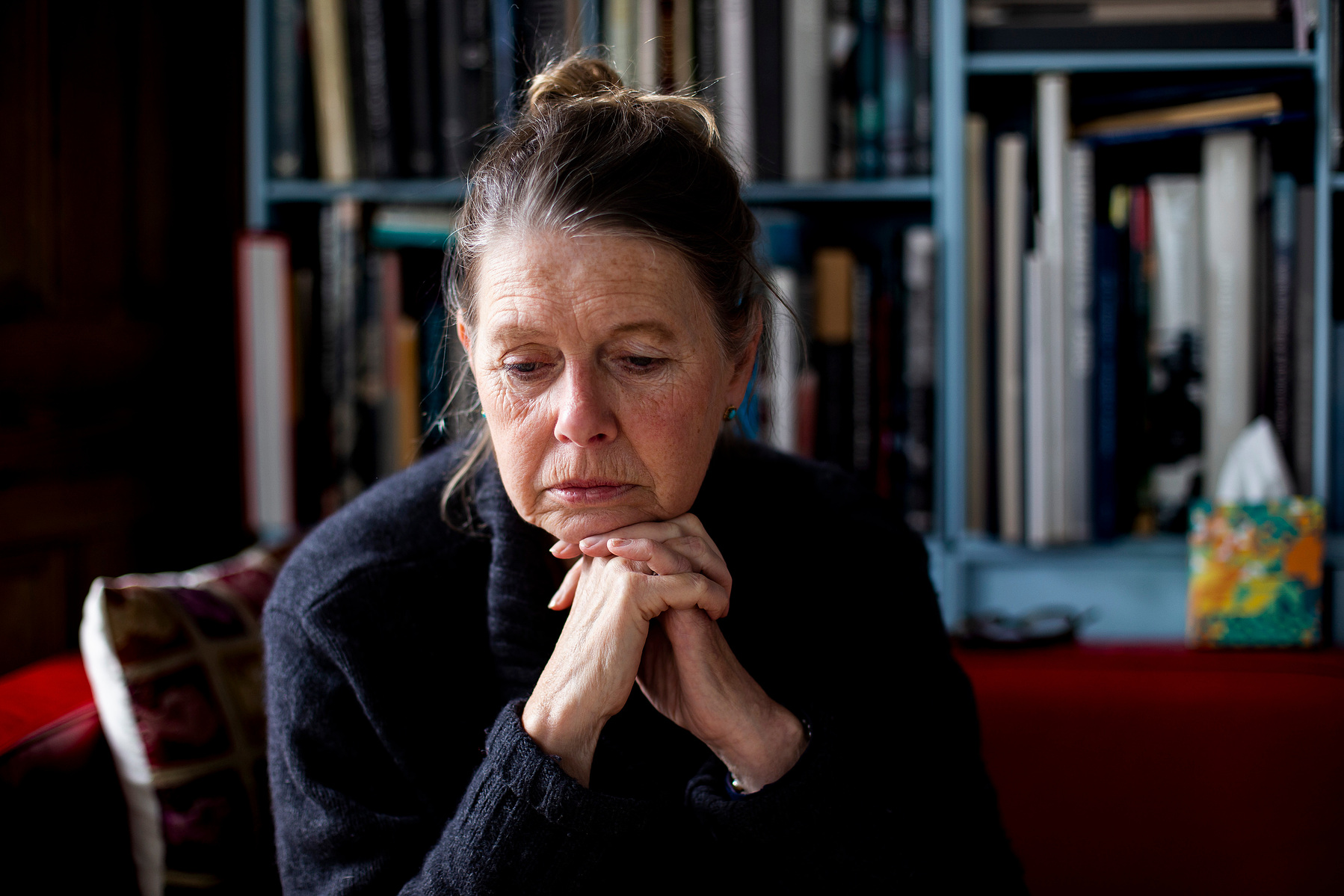 Zelda Cheatle, sits forward on a red leather sofa, her chin resting on her interlocked fingers, her eyes to the ground.  Light strikes her face from the left of the image, while behind and to her right are somewhat darker. A bookcase is in the background.