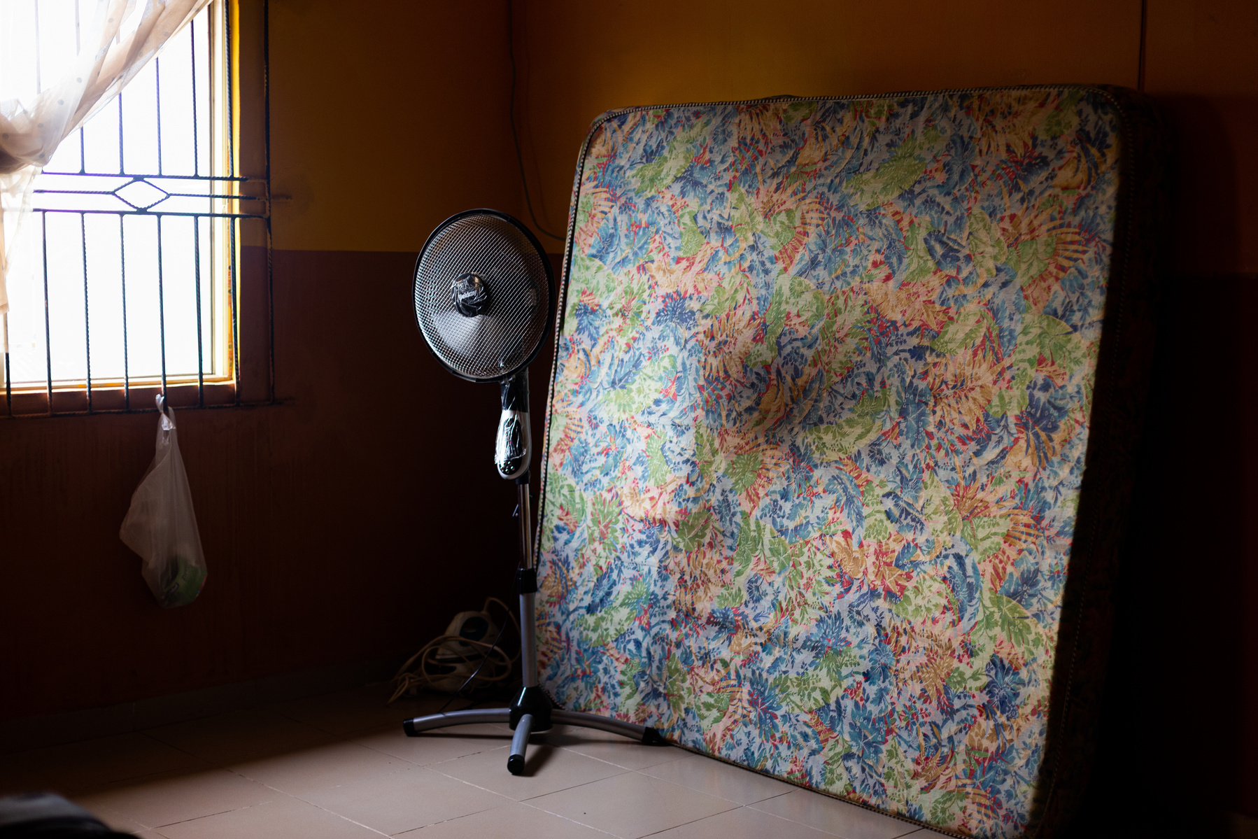 The corner of a bedroom - on the left, a window covered in iron bars, and a net curtain pulled to one side; on the right, a floral double mattress stands against the wall.  A standing fan is set in front of the mattress, it is not switched on.