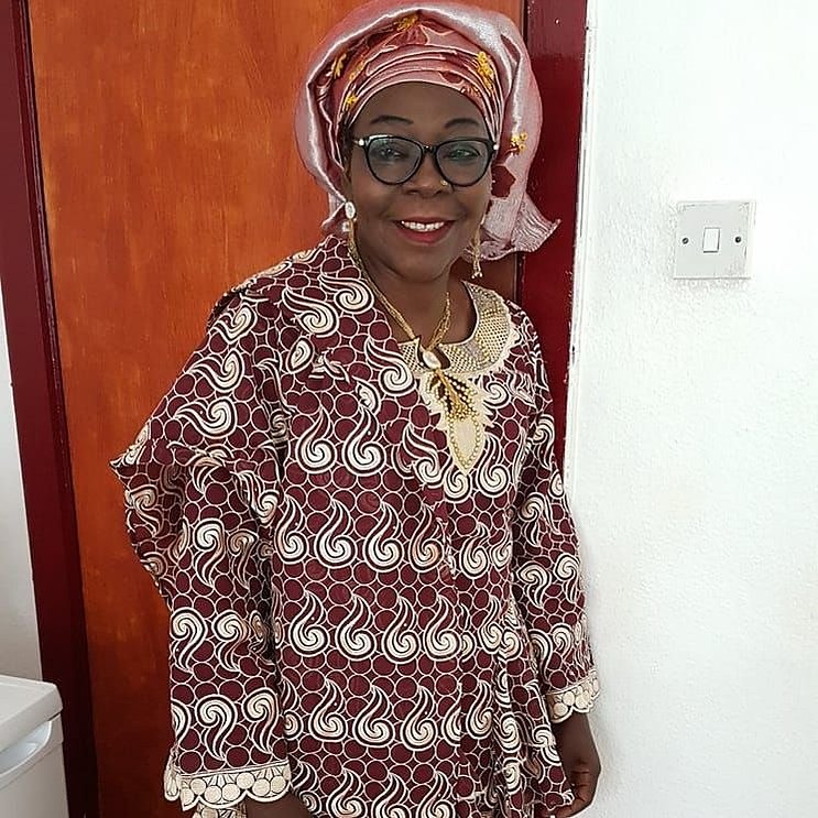 My mum, dressed in a maroon and gold outfit with a deep red and gold headtie, and a gold necklace and earrings, stands in front of a bedroom door, looking slightly off camera with a slight smile on her face.