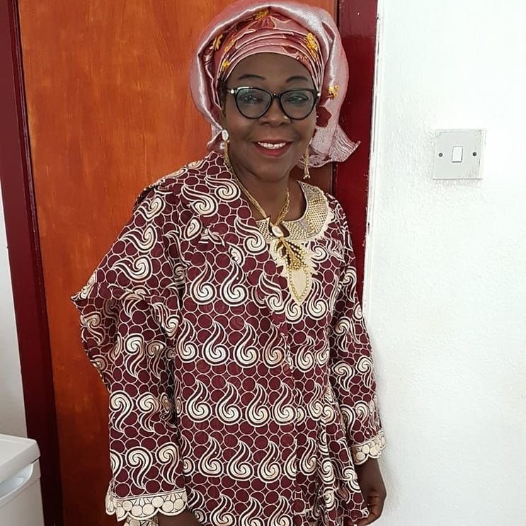 My mum, dressed in a maroon and gold outfit with a deep red and gold headtie, and a gold necklace and earrings, stands in front of a bedroom door, looking slightly off camera with a slight smile on her face.