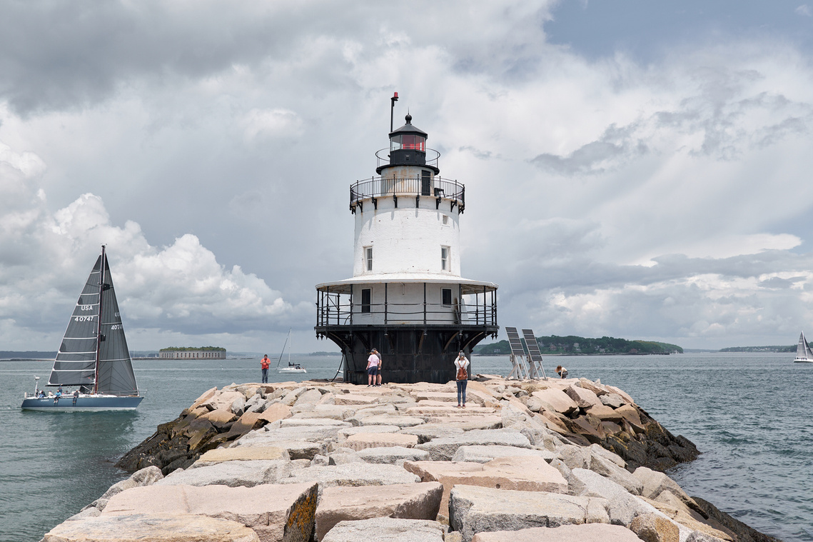 Spring Point Ledge Lighthouse in Portland, Maine with Fort Gorges in the background as a sailboat passes by.