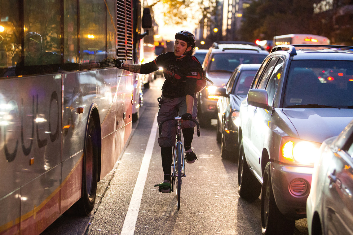 Bike messenger rides between lanes of traffic while running his hand along a moving bus.