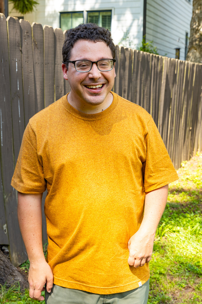 A young man with brown hair and glasses smiles as he stands outside. 