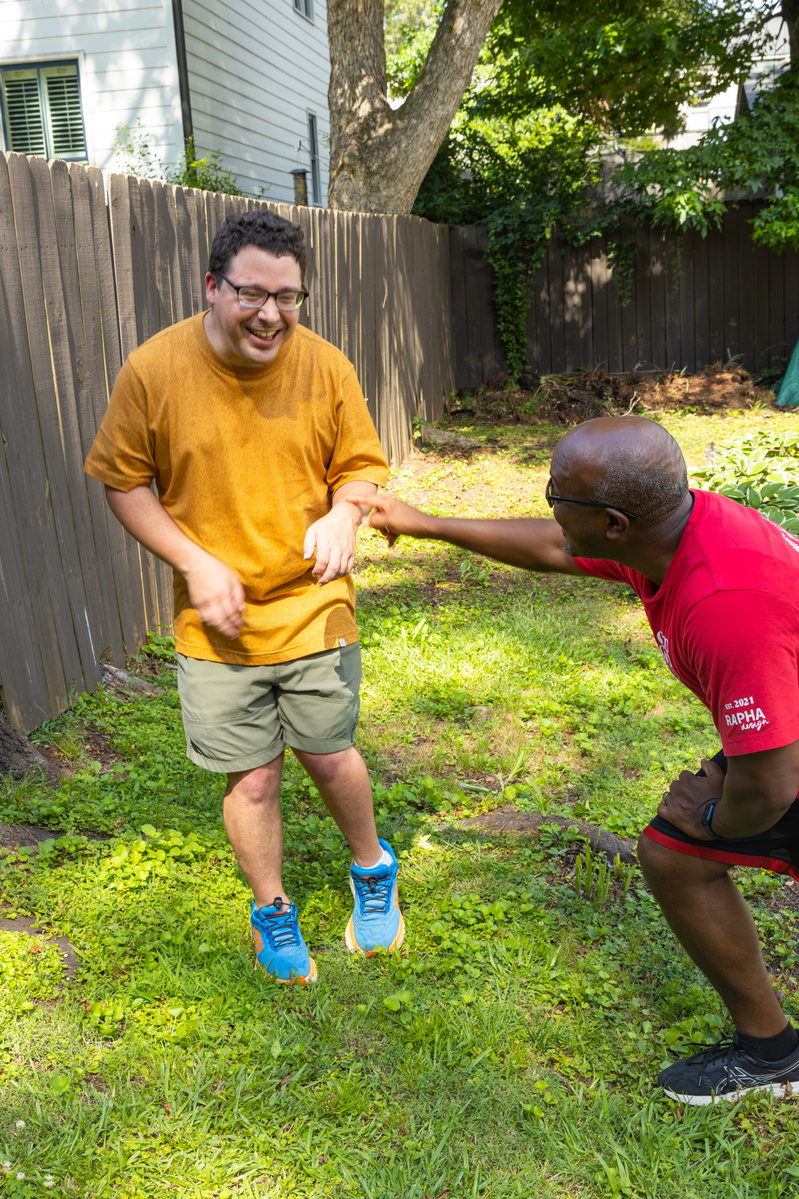 Two men laugh as they stand outside in front of a wooden fence.