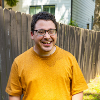 A young man with brown hair and glasses smiles as he stands outside. 